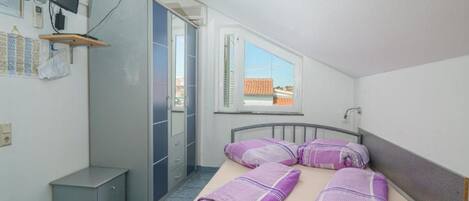 Une petite pièce propre et bien rangée, avec un lit, un bureau, une chaise, une armoire, un téléviseur et une fenêtre donnant sur des immeubles.