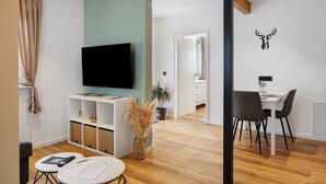 A modern living room with a flat-screen TV, a dining area with a table and chairs, and a hallway leading to a bathroom.