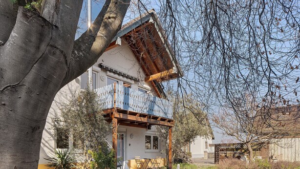 A two-story house with a balcony, a garden, and a tree in the foreground.