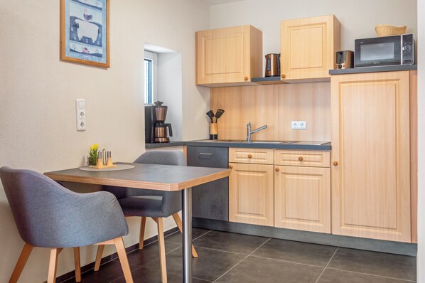 Cocina compacta con armarios de madera, cafetera y microondas. Hay una mesa de comedor con sillas.