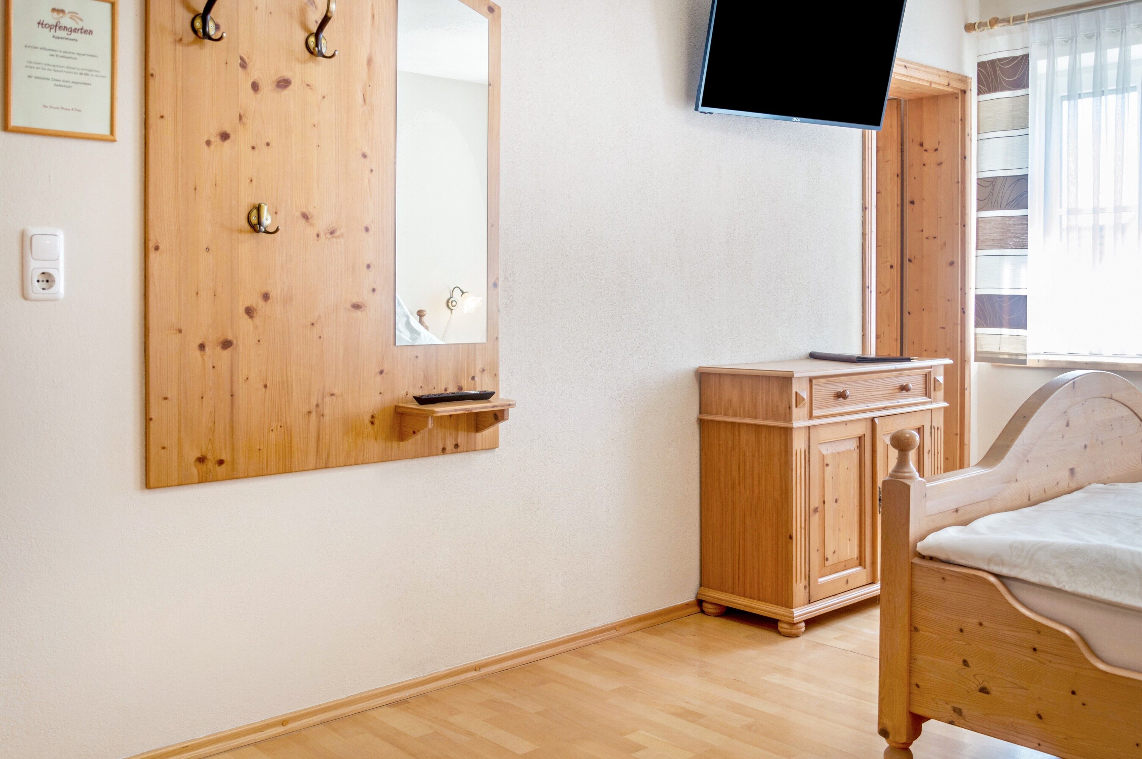 A bedroom with a wooden wardrobe, a bed, a television mounted on the wall, and a window with curtains.