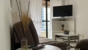 Ett hotellrum med en säng, en tv som är monterad på väggen, ett litet bord med en vas och fjärrkontroller samt ett fönster med gardiner.