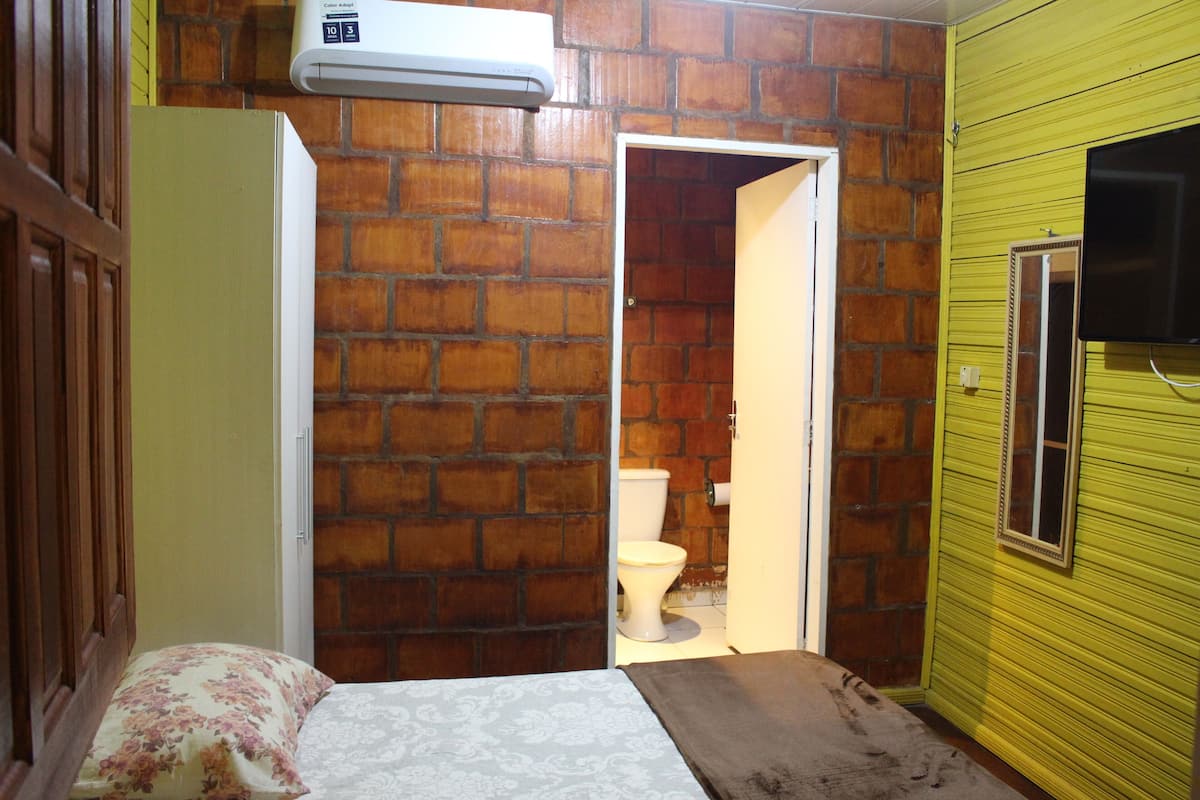 Una habitación con pared de ladrillo, una cama con almohada estampada, un inodoro blanco y un televisor instalado en la pared amarilla.