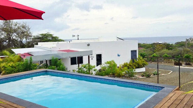 A swimming pool with a wooden deck, a red umbrella, and a white building with a red roof in the background. - Villa Aloë - Ocean view villa with pool in Lagun (Lagun)
