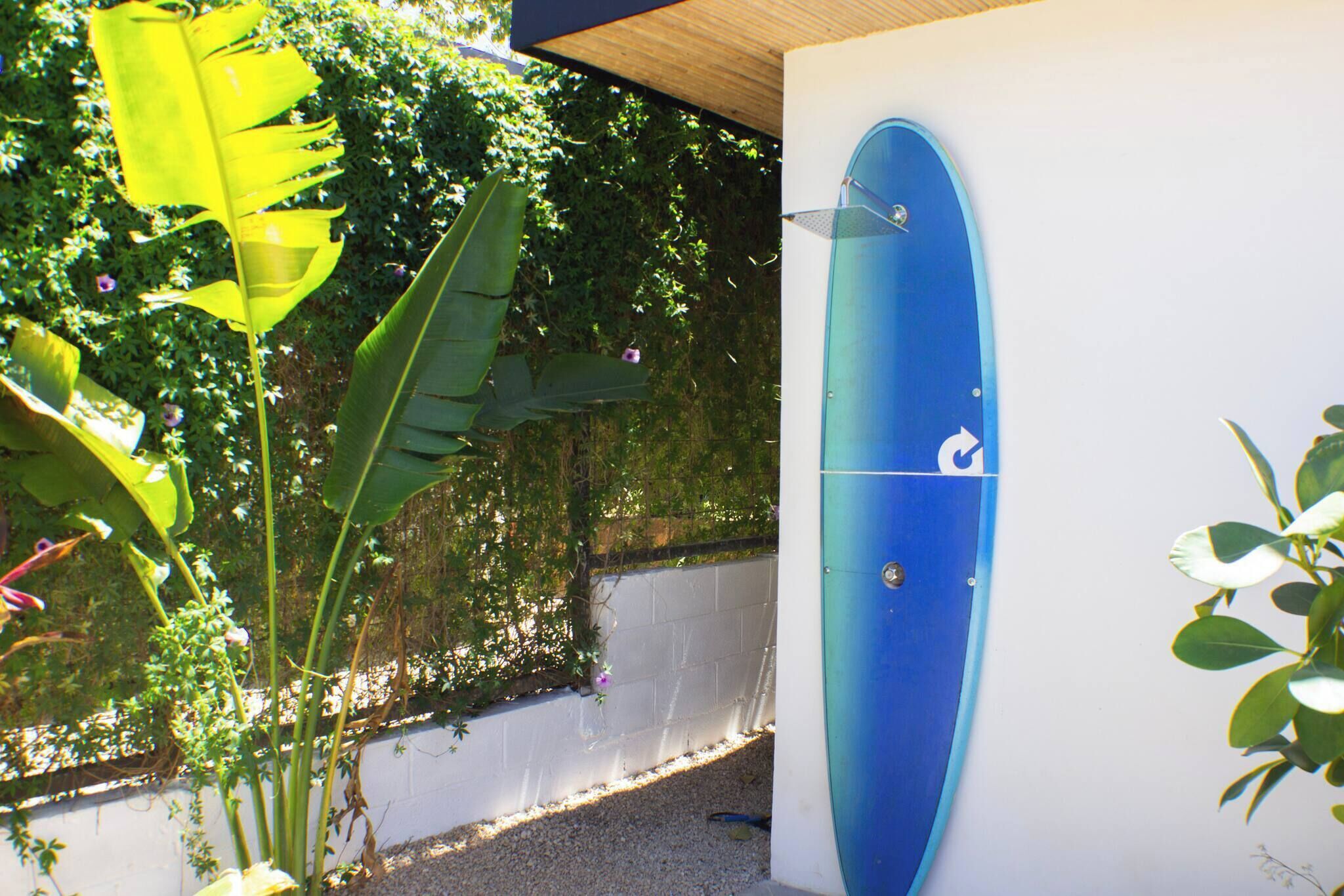 Une planche de surf bleue est fixée au mur, entourée de verdure et adossée à un mur blanc.