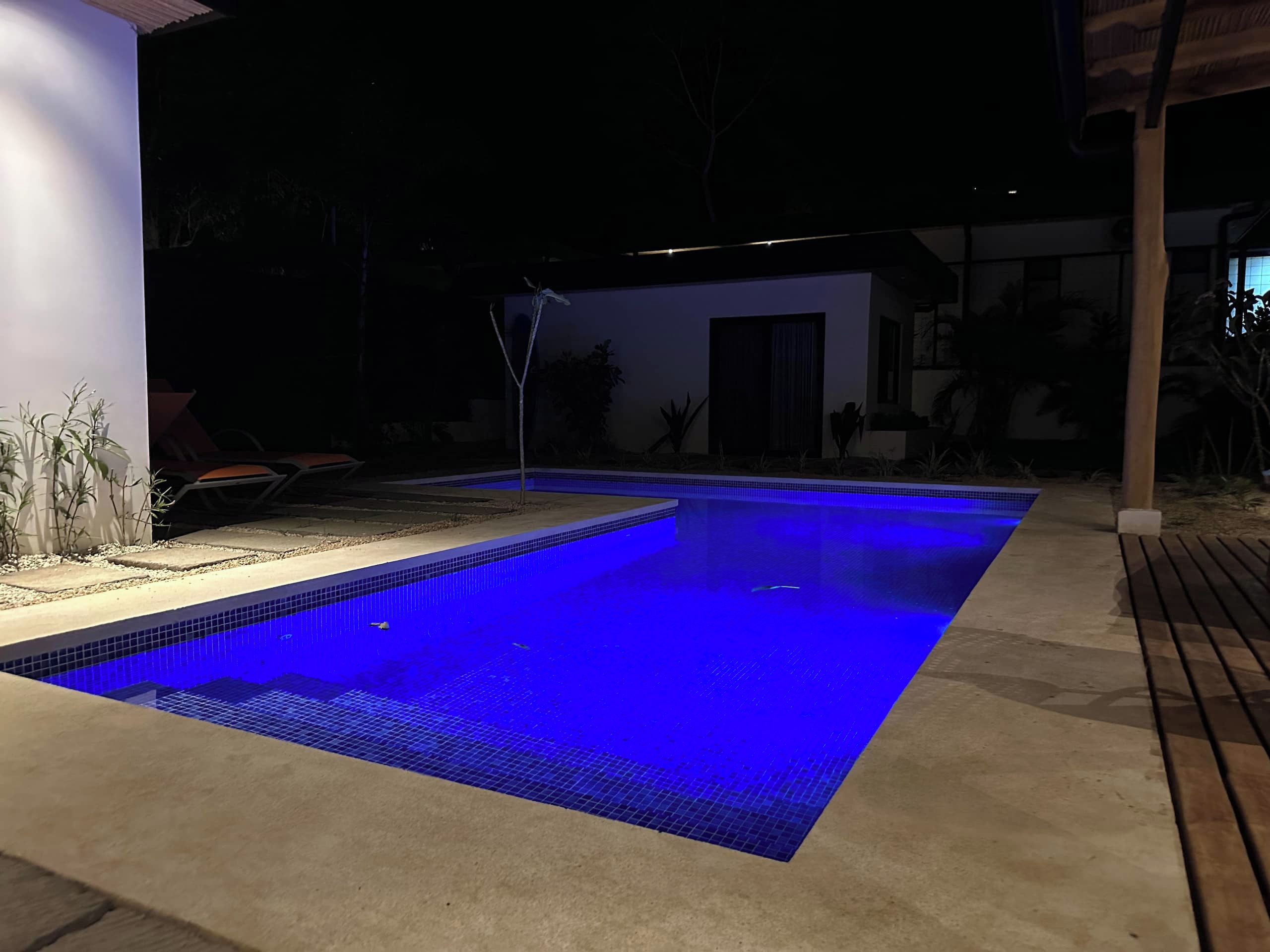 A rectangular swimming pool with a tiled edge and a blue-lit interior, surrounded by a concrete deck and a covered area with chairs.