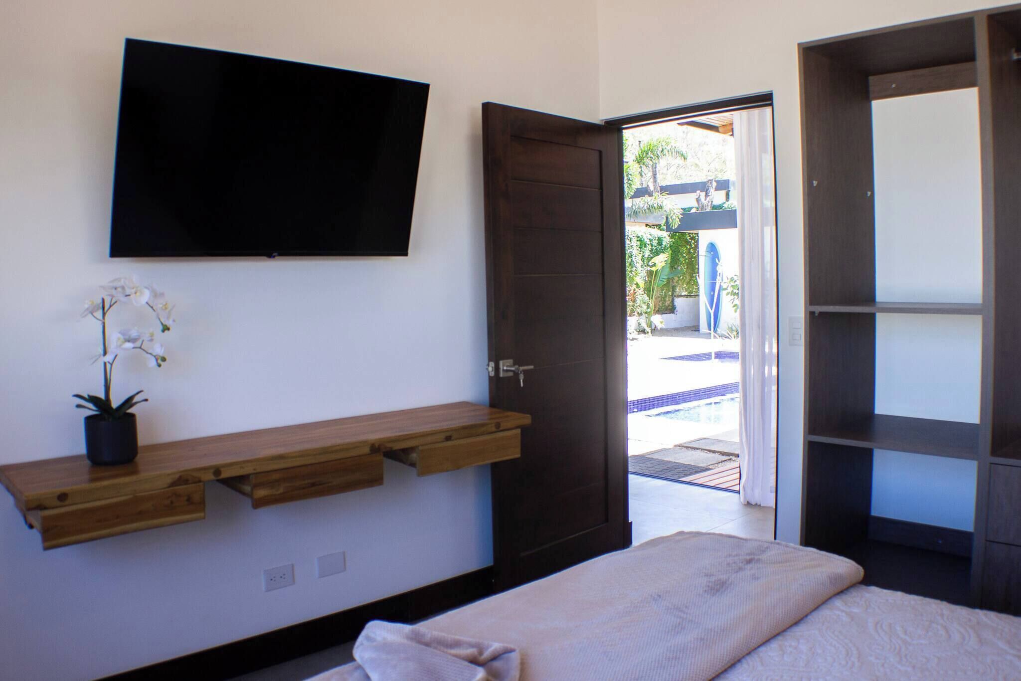 A bedroom with a bed, a mounted TV, a wooden shelf with a potted orchid, and a door leading to an outdoor area.