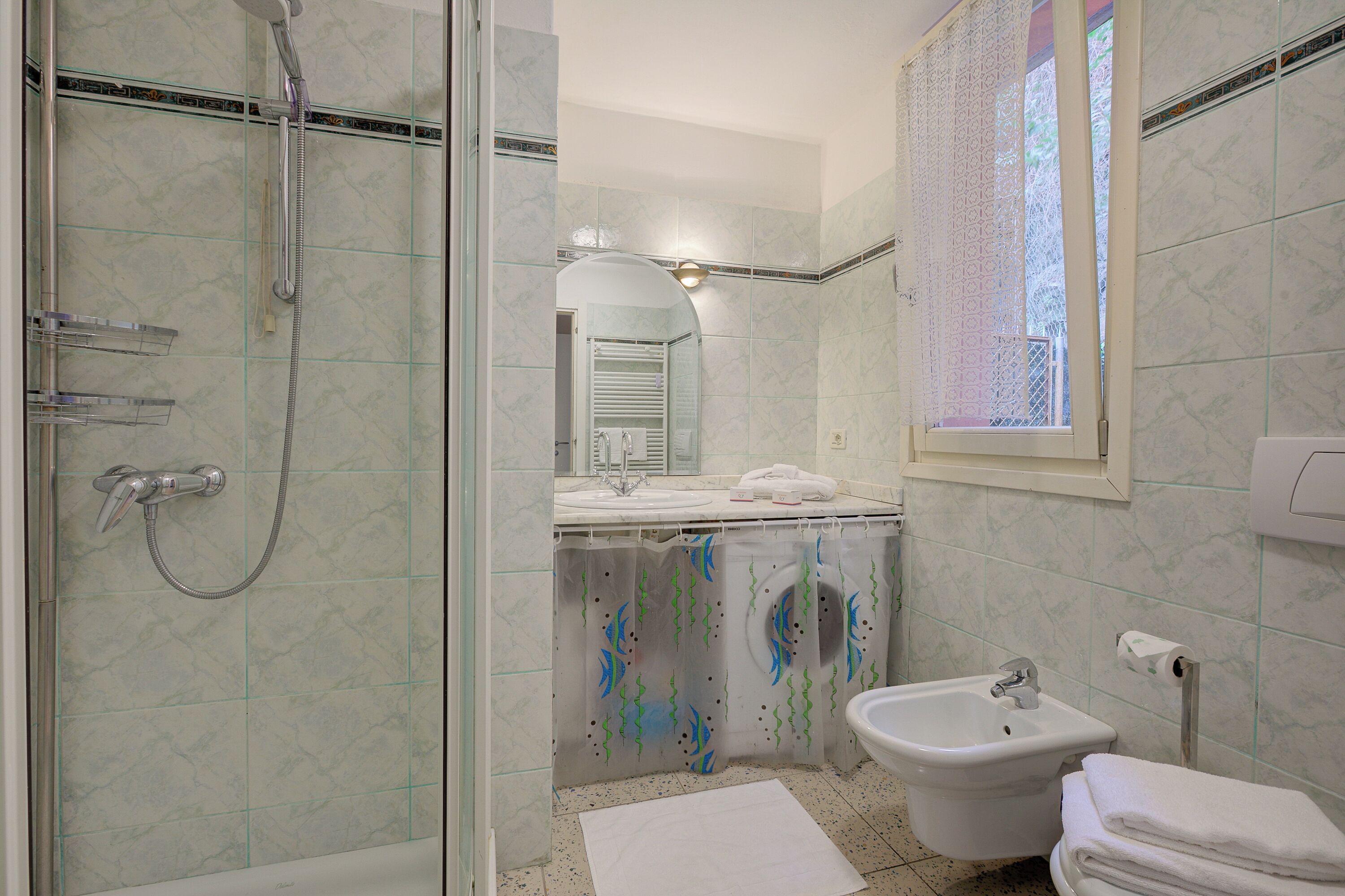 A bathroom with a shower, sink, bidet, and a window with a view.