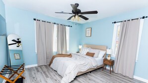 Ein Schlafzimmer mit einem Bett, Deckenventilator, Nachttisch, Lampe und einem Fenster mit Vorhängen.
