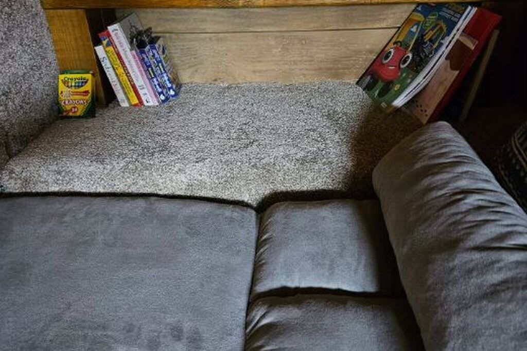 Un canapé-lit recouvert d’un tissu gris, doté d’une tête de lit en bois et d’une étagère où sont posés des livres et une boîte de crayons de couleur.