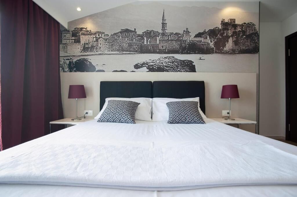 A hotel room with a large bed, two bedside lamps, a headboard, and a cityscape mural on the wall.