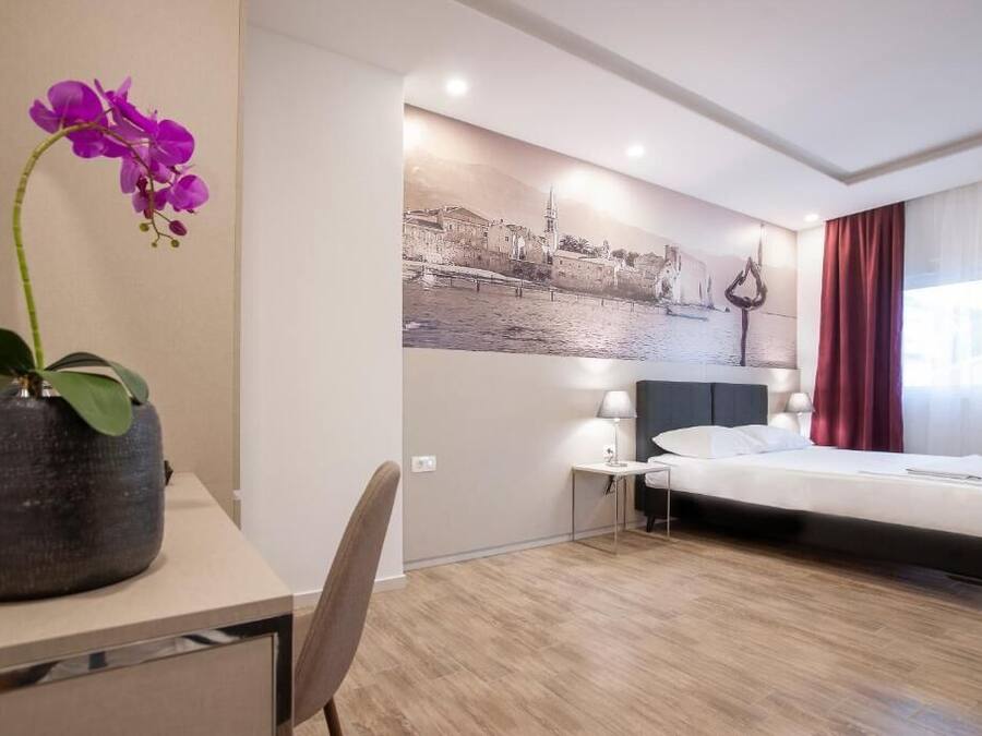 Ein Hotelzimmer mit einem Bett, einem Schreibtisch mit Blumen, einem Stuhl, einem Nachttisch mit einer Lampe und einer Wand mit einem Stadtbild.