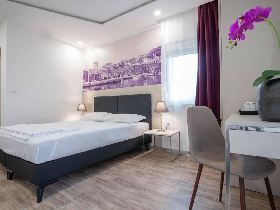 Una habitación de hotel con cama, mesitas de noche, escritorio, una silla y una orquídea morada.