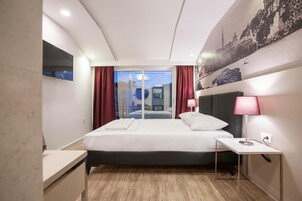Habitación de hotel con cama, mesita de noche, lámpara y vistas a través de un ventanal.