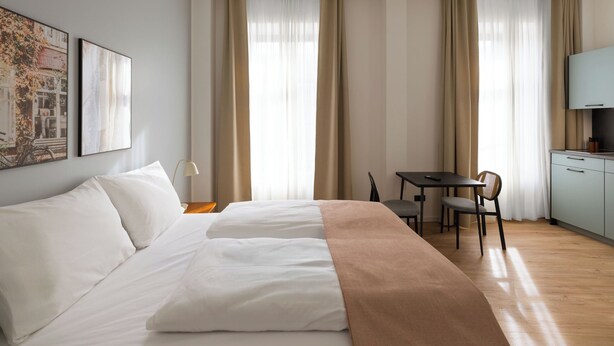 Quarto de hotel com cama, escrivaninha, cadeira e cozinha compacta.