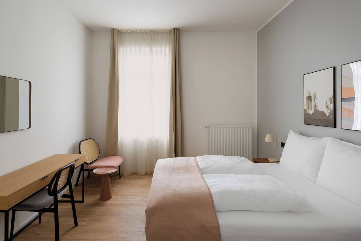 Habitación de hotel con cama, escritorio, silla, radiador y ventana con cortinas.