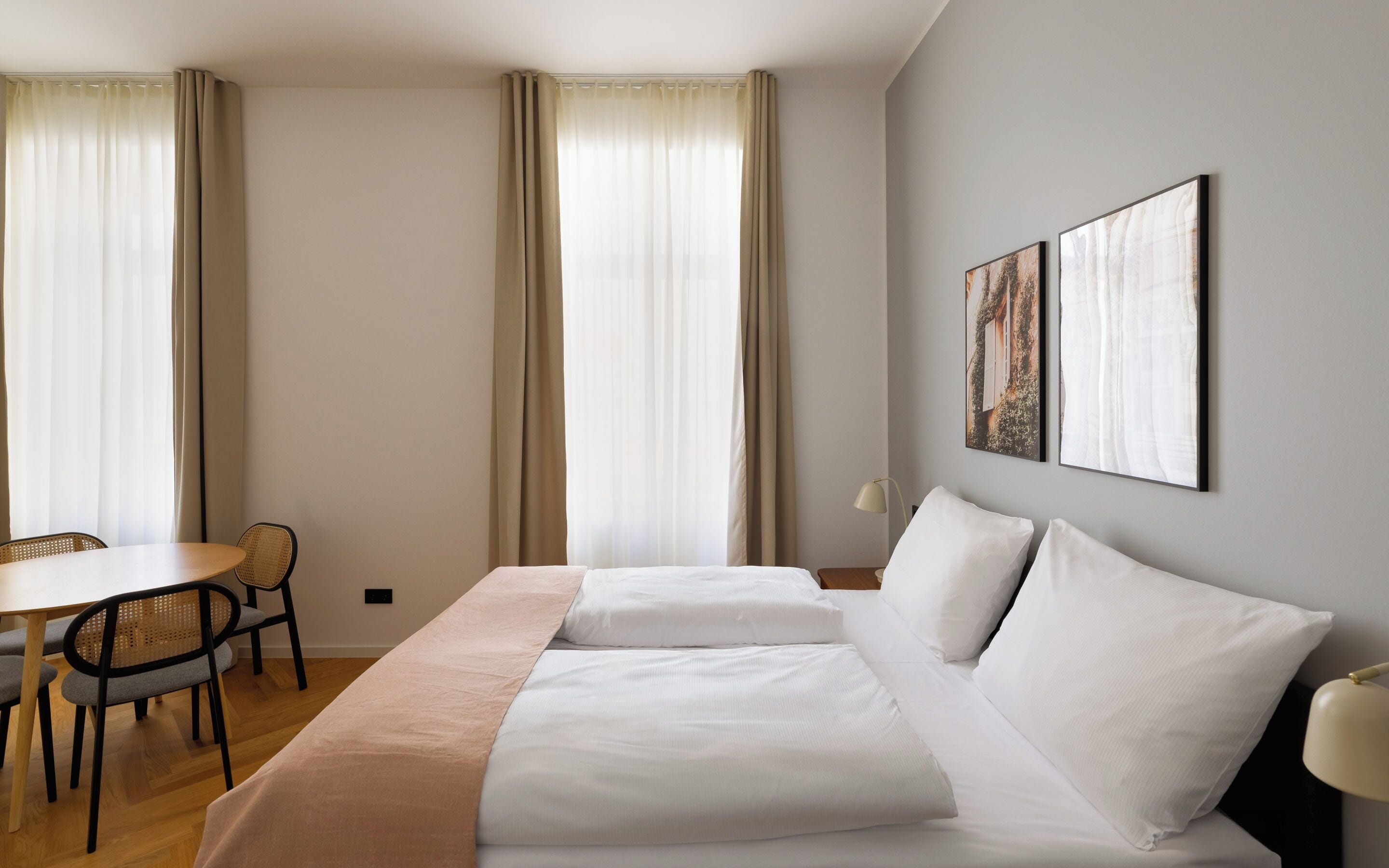 Una habitación de hotel con cama, escritorio, dos sillas y dos ventanas con cortinas.