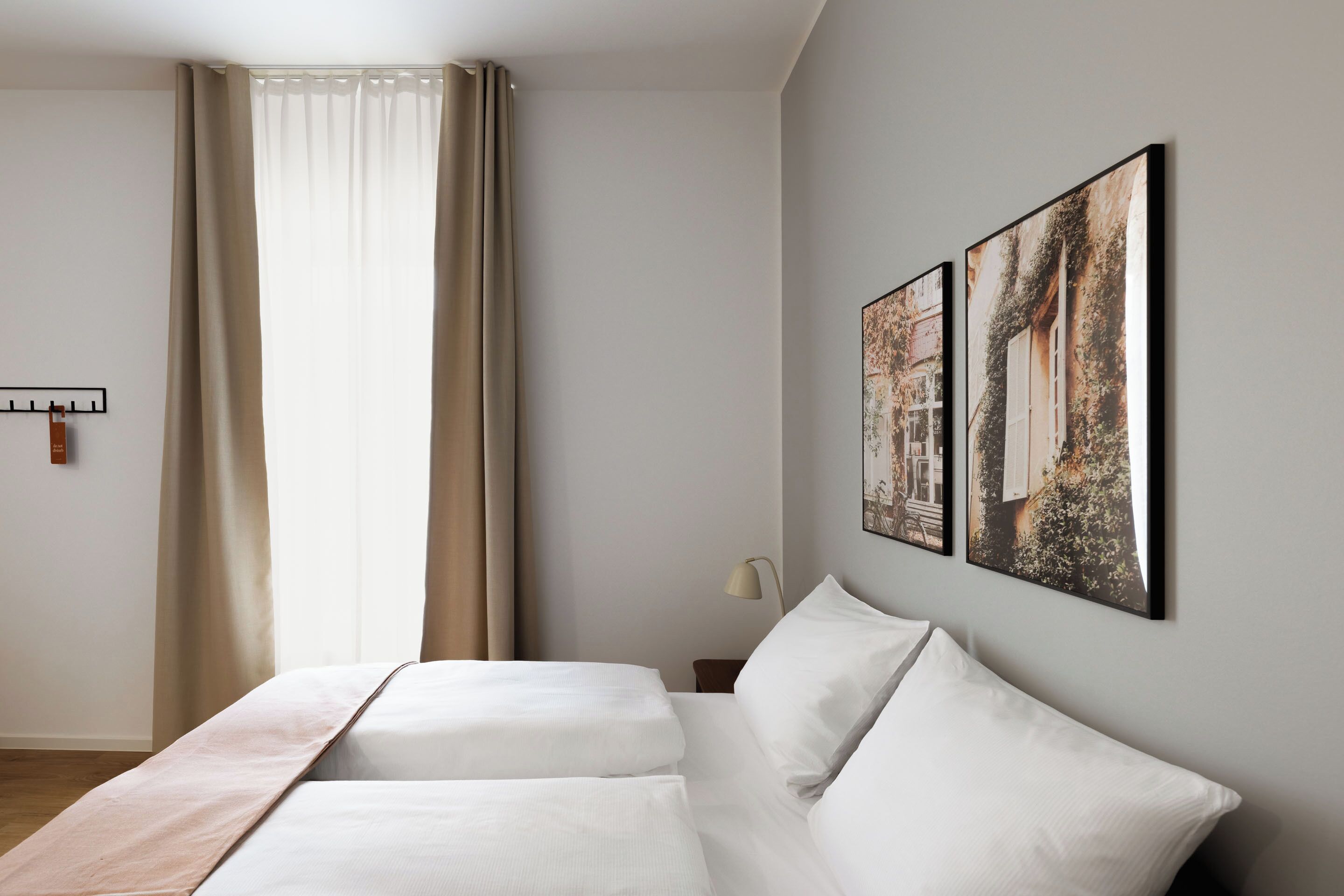 Ein Hotelzimmer mit Bett, Vorhängen, einem Nachttisch, einer Lampe und zwei gerahmten Bildern an der Wand.