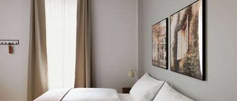 Habitación de hotel con cama, cortinas, mesita de noche, lámpara y dos cuadros en la pared.