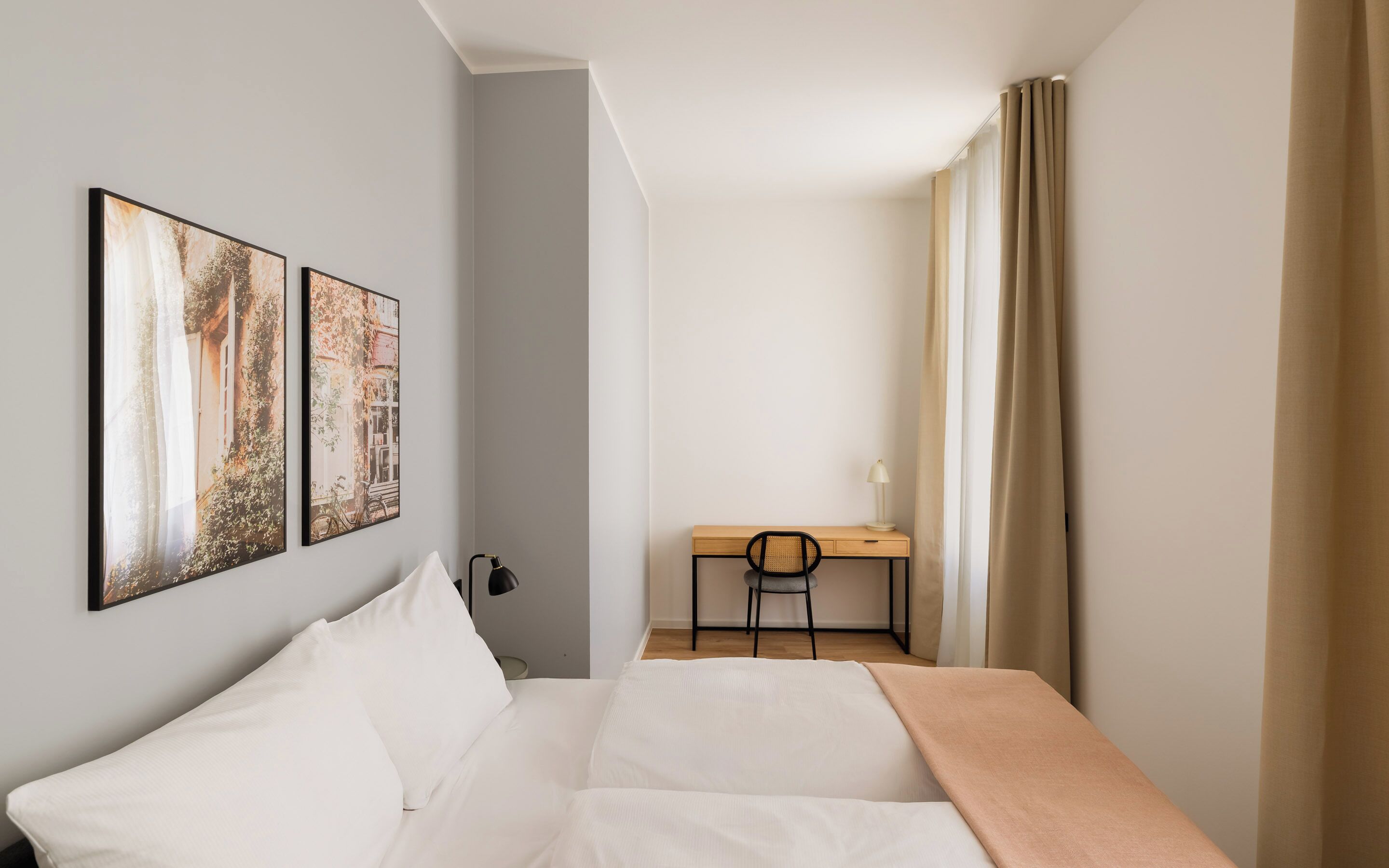 A hotel room with a bed, a desk, a chair, and two framed pictures on the wall.