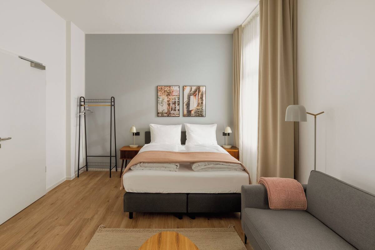 Una habitación de hotel moderna con cama, mesitas de noche, sofá, lámpara de pie y zona de ducha.