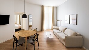 Uma sala de estar moderna com uma mesa de jantar redonda de madeira, duas cadeiras cinzas estofadas, um sofá bege, uma TV de tela plana na parede, um abajur e um espelho grande.