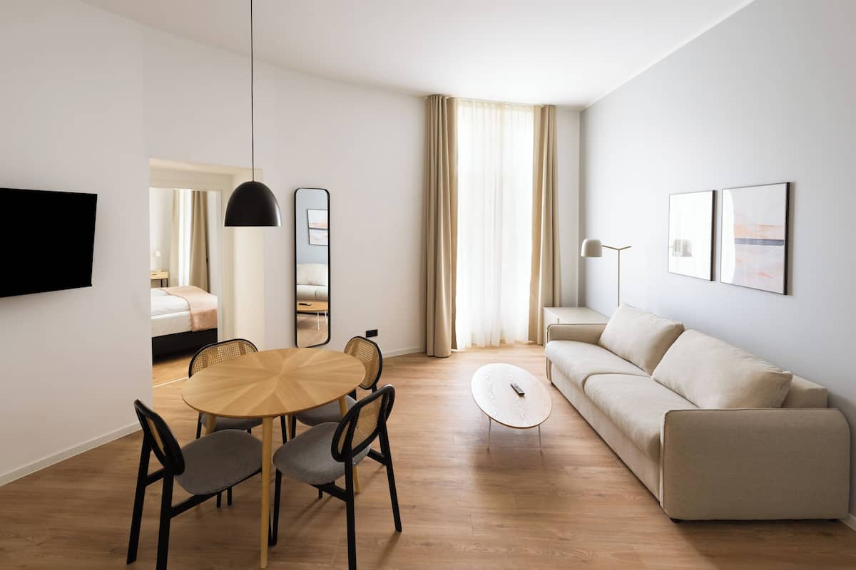 Una sala moderna con una mesa de comedor redonda de madera, dos sillas tapizadas de gris, un sofá beige, un televisor de pantalla plana montado en la pared, una lámpara de pie y un espejo grande.