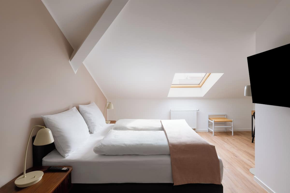 Un dormitorio con tragaluz, una cama con almohadas, una mesita de noche con lámpara, una silla y un televisor.