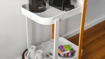 A white, three-tiered, wheeled coffee cart with a black coffee maker and a black electric kettle on the top shelf, a tray with cups and spoons on the middle shelf, and a tray with tea packets and a small toy on the bottom shelf.