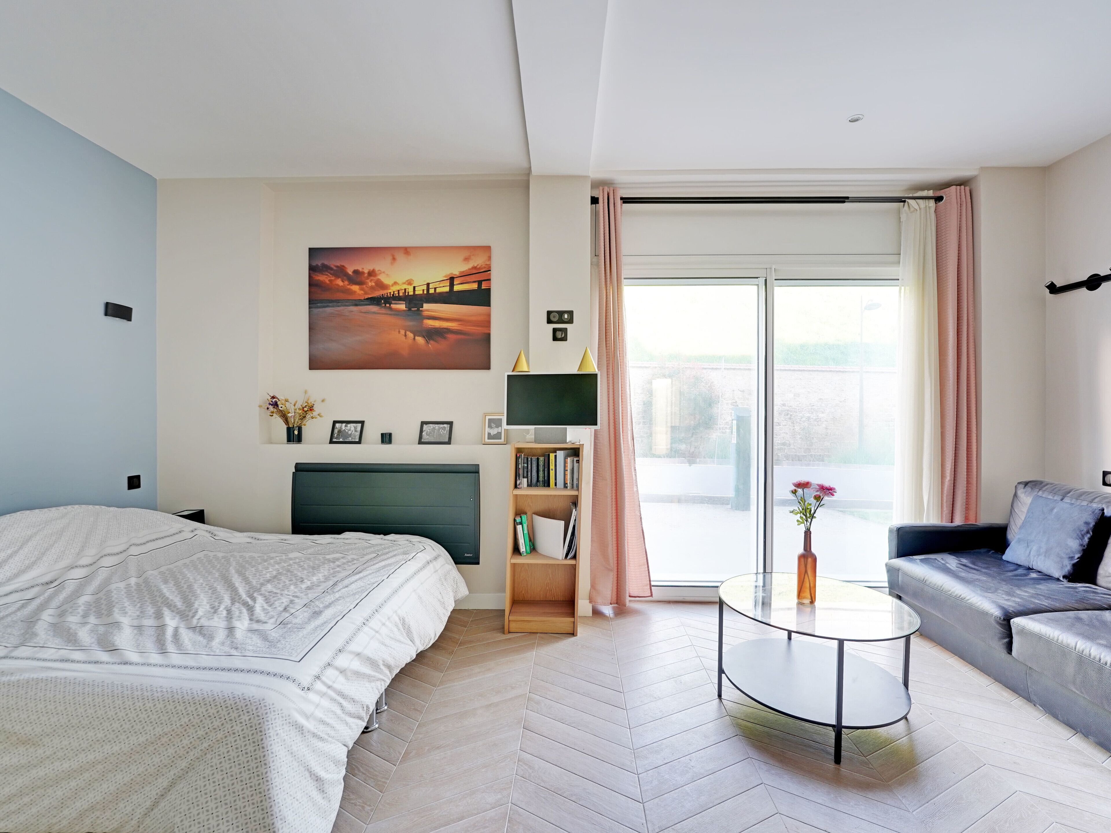 Une chambre à coucher moderne avec un lit, un canapé, une télévision, une bibliothèque et un tableau représentant un coucher de soleil.