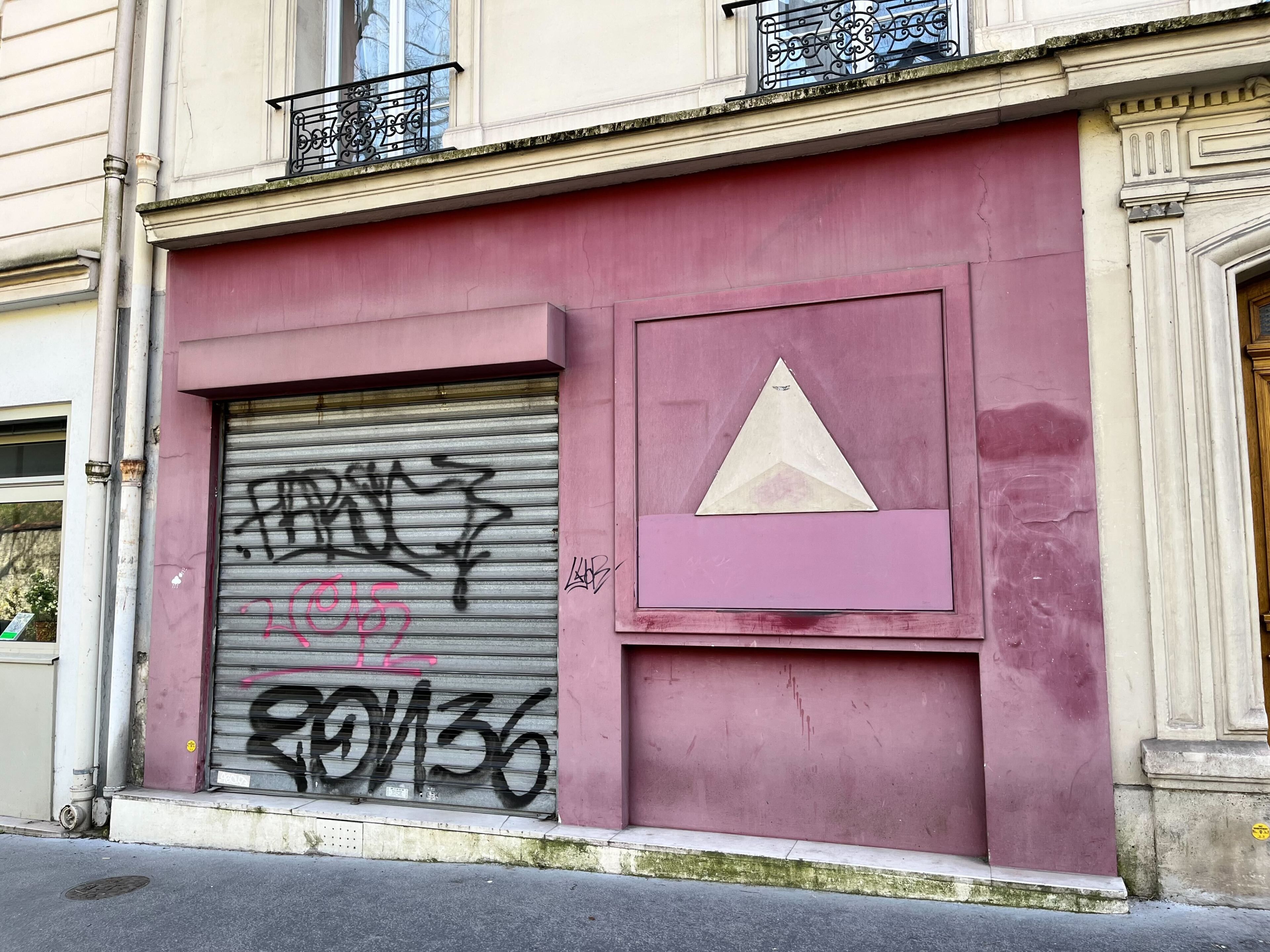 Une façade d’immeuble avec un volet fermé, des graffitis et un élément architectural triangulaire.