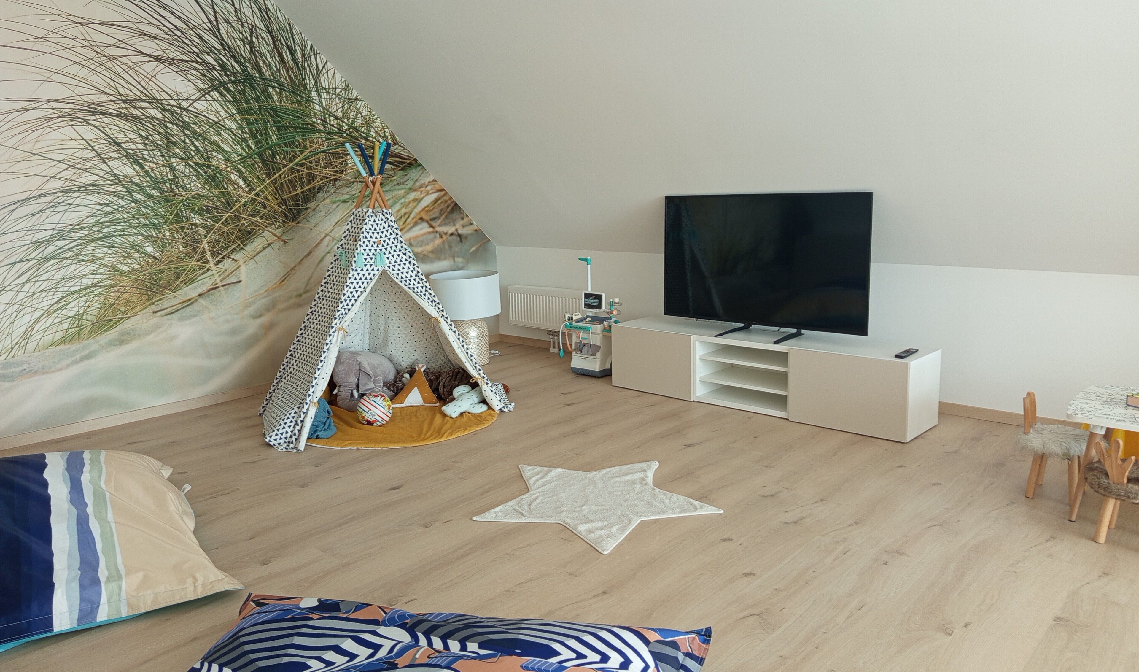Een woonkamer met een televisie, een witte entertainmentkast, een stervormig vloerkleed, een tent en een grote plant.