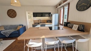 Ein kompakter Wohnraum mit Küchenzeile, einer Essecke mit Holztisch und weißen Stühlen, einem blauen Sofa und einer großen Wanduhr.