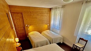 A room with two beds, a wooden wall, a small table with a lamp, and a window with curtains. - Spacious apartment in village center, shops 2 steps away. (Val-Cenis)