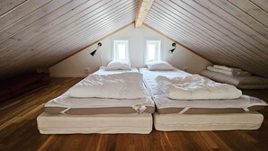 A room with two beds, wooden flooring, a sloped ceiling, and two windows.
