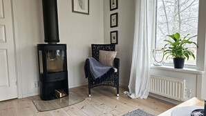 A cozy room with a wood-burning stove, a wicker chair, a potted plant, and framed pictures on the wall.
