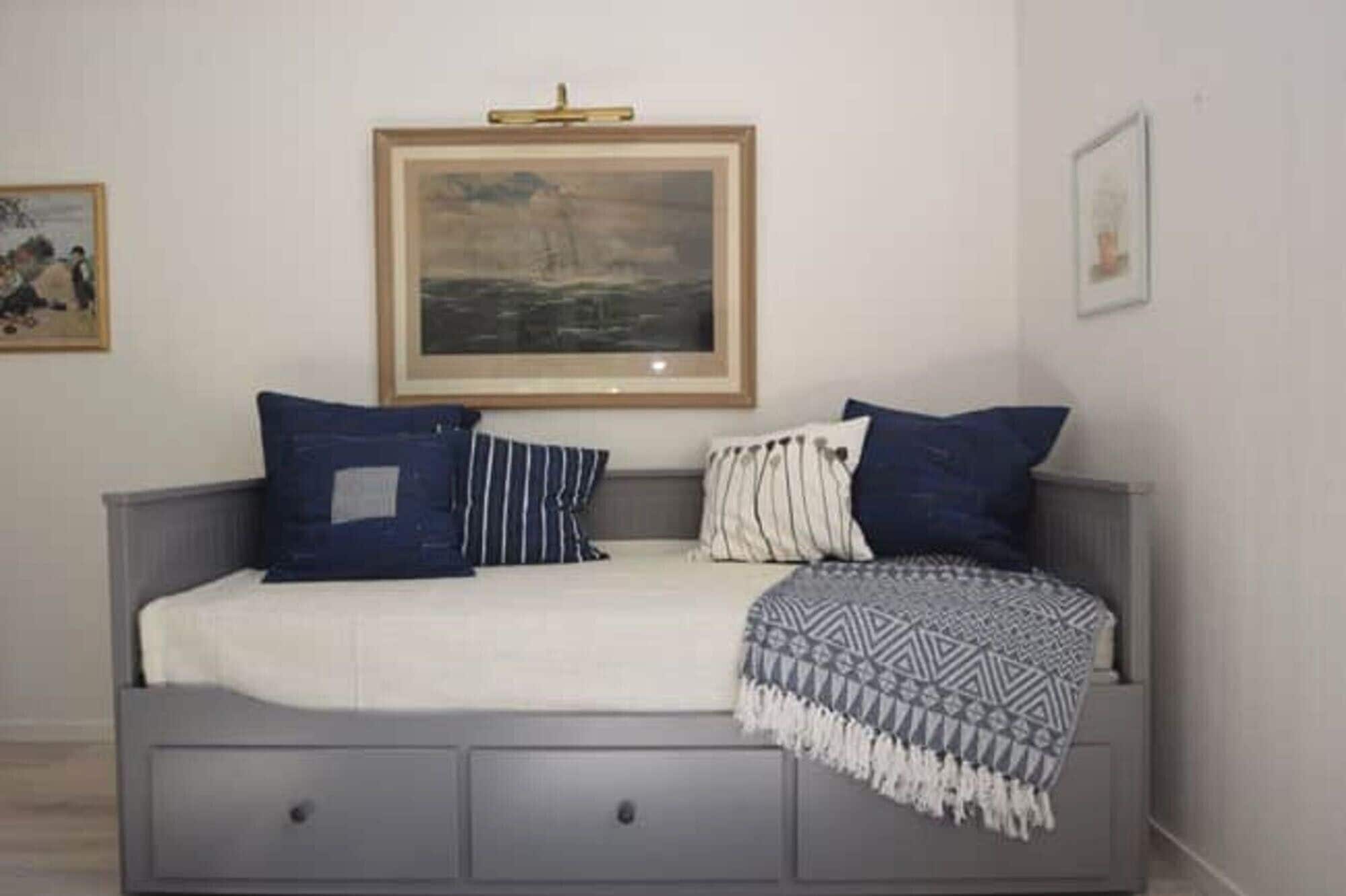 A room with a grey sofa, a framed painting above it, and a small table with drawers.