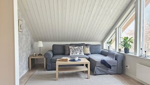 Um quarto estilo loft aconchegante com teto inclinado, um sofá cinza, uma mesa de centro de madeira e uma grande janela com plantas.