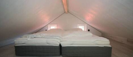 Un dormitorio ático pequeño y triangular con una cama individual, techo inclinado y dos ventanas pequeñas.