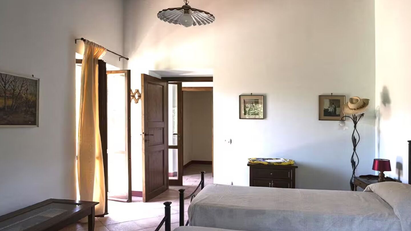 A bedroom with a bed, a bedside table, a lamp, a wardrobe, and a door leading to another room.