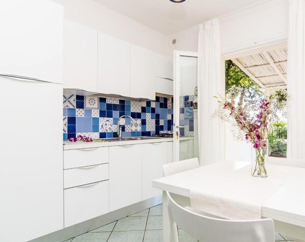 A kitchen with white cabinets, a blue and white tiled backsplash, and a dining area with a white table and chairs. - Colibrì2 by ColombiElbaVacanze (Capoliveri)