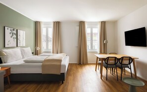 A hotel room with a bed, a desk, a chair, a TV, and two windows with curtains. - Limehome Wels Ringstraße (Wels)