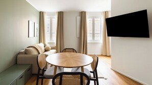 A modern living room with a sofa, round dining table, and chairs, a TV mounted on the wall, and large windows with curtains. - Limehome Wels Old Town (Wels)