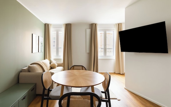 A modern living room with a sofa, round dining table, and chairs, a TV mounted on the wall, and large windows with curtains. - Limehome Wels Old Town (Wels)