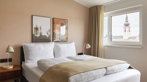 A hotel room with a bed, two bedside lamps, a window with a view of a tower, and two framed pictures on the wall. - Limehome Wels Old Town (Wels)