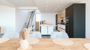 A modern kitchen with a central island, a dining area with a wooden table and chairs, and a staircase leading to an upper level. - Villa Bellevue (Donville-les-Bains)