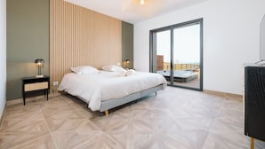 A bedroom with a bed, bedside tables, a lamp, a large window, and a view of the outside. - Villa Bellevue (Donville-les-Bains)