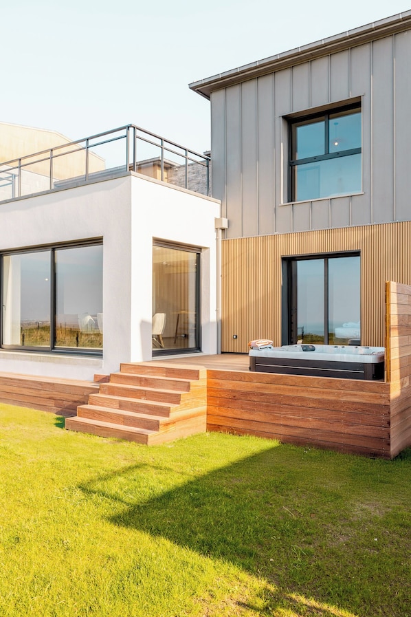 A modern two-story house with large windows, a hot tub, and a wooden deck.