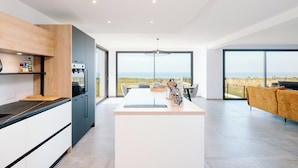 A modern kitchen with a large island, a dining area with a view, and a living space with a sofa. - Villa Bellevue (Donville-les-Bains)