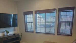 A living room with a flat-screen TV, a brown leather sofa, a wooden coffee table, and three windows with blinds.